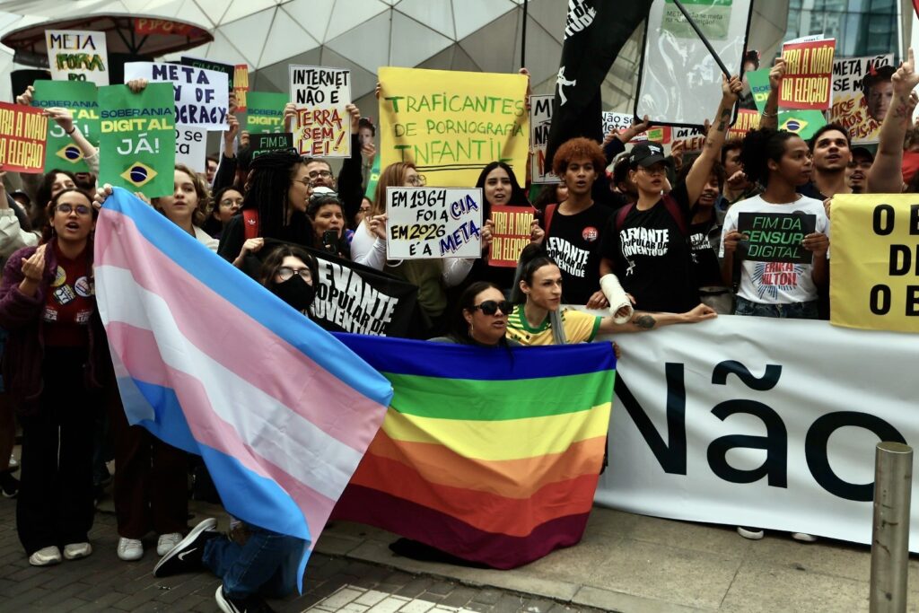 Manifestantes seguram bandeiras LGBT e trans, além de cartazes com frases como "Soberania Digital Já!" e "Tira a mão da eleição" em protesto em frente à sede da Meta.