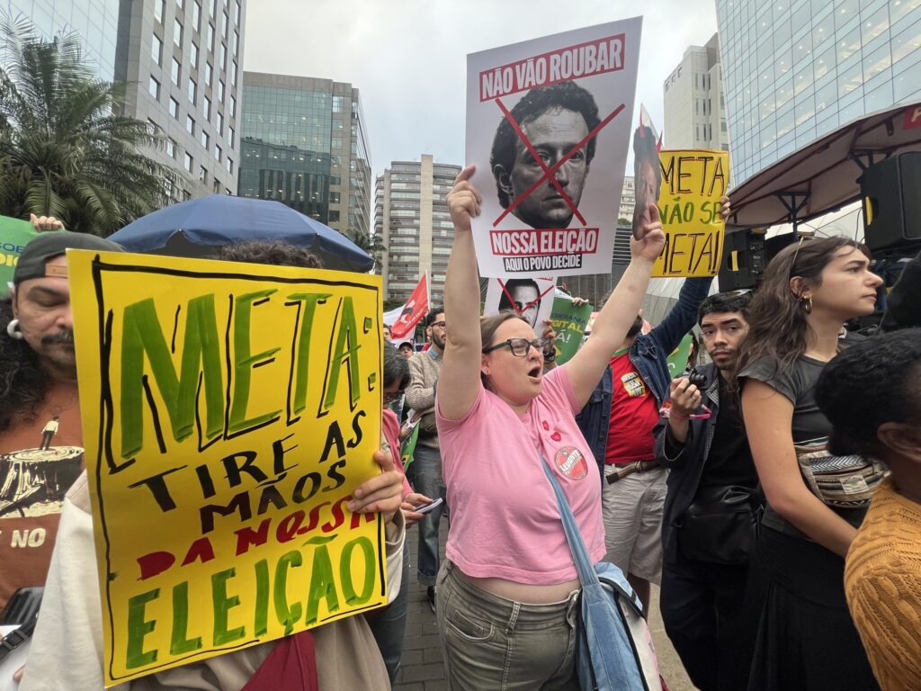 falhas de moderação nas eleições: Manifestantes seguram cartazes com os dizeres "META: TIRE AS MÃOS DA NOSSA ELEIÇÃO" em frente a prédios comerciais.