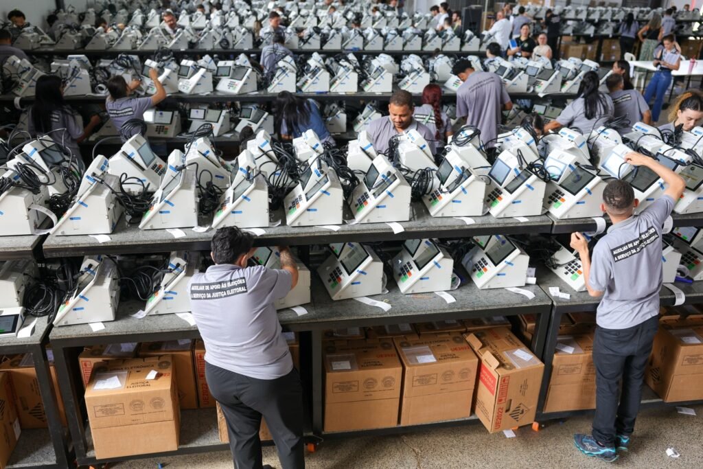 esplanada dos ministerios: Técnicos preparam fileiras de urnas eletrônicas sobre bancadas em um galpão, com caixas de papelão da Justiça Eleitoral.
