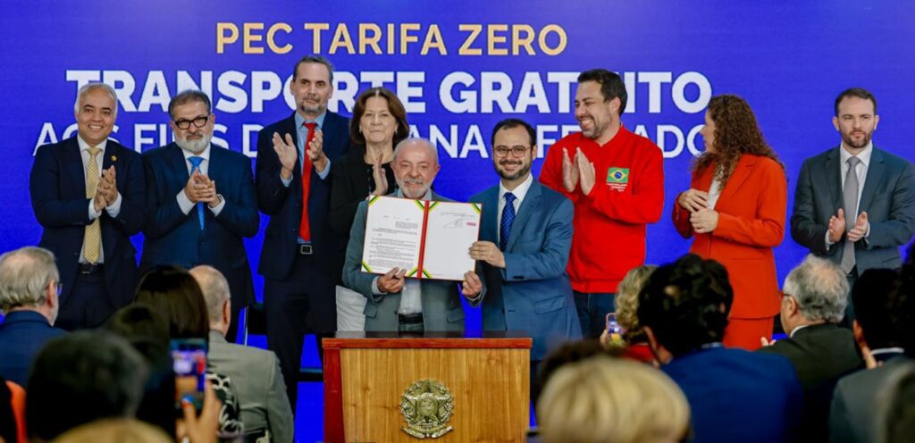 Presidente Luiz Inácio Lula da Silva segura um documento diante de um painel azul com os dizeres "PEC TARIFA ZERO" e "TRANSPORTE GRATUITO", cercado por pessoas que aplaudem.