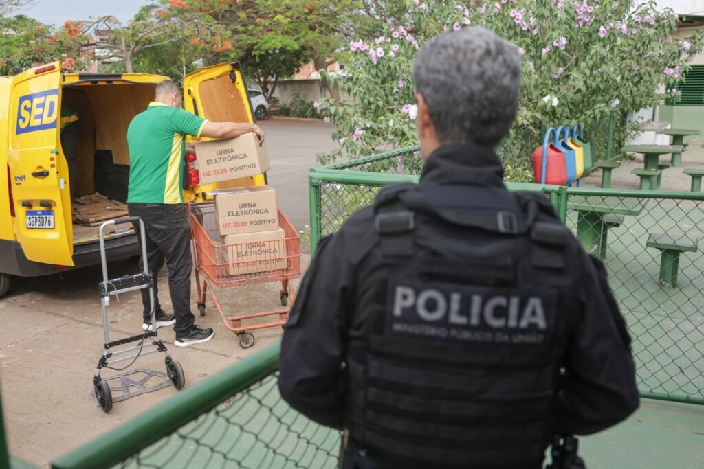 assédio eleitoral: Homem fardado de costas observa funcionário descarregando caixas de urnas eletrônicas.