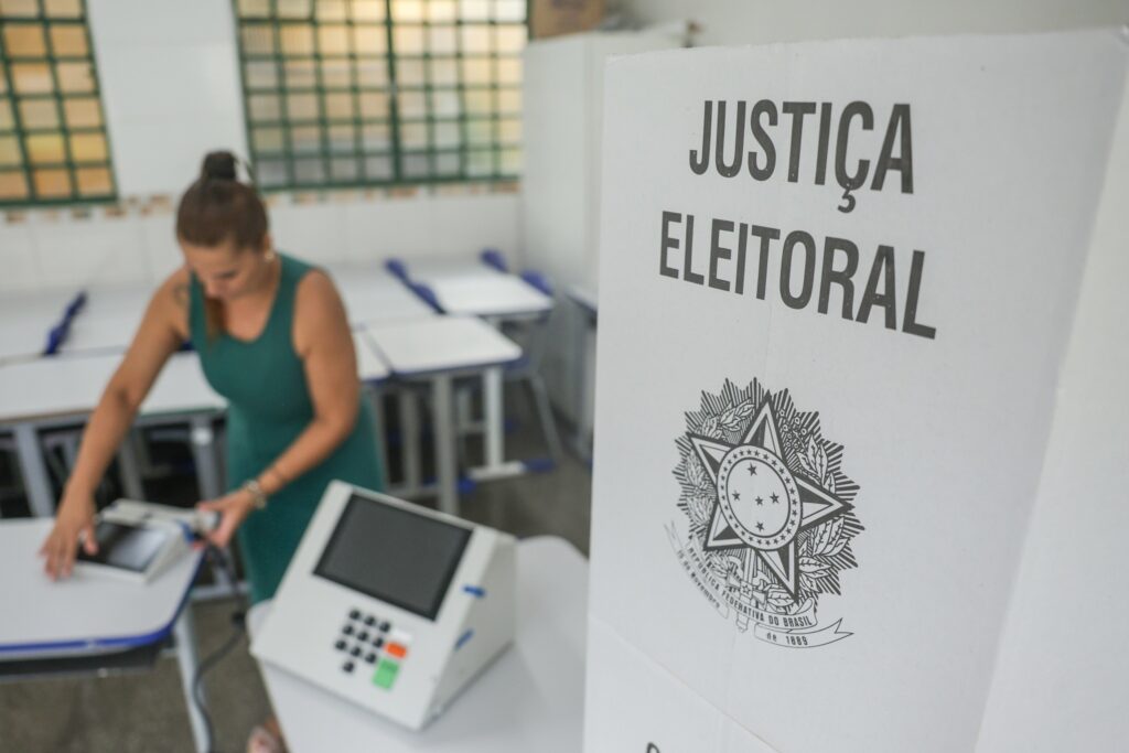 Mulher prepara urnas eletrônicas em sala de aula, com um painel em primeiro plano com os dizeres "JUSTIÇA ELEITORAL".