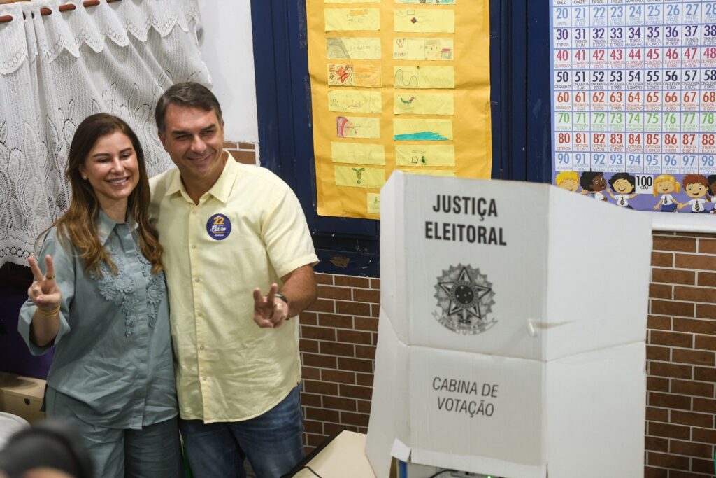 O senador Flávio Bolsonaro e uma mulher posam sorridentes ao lado de uma cabina de votação de papelão com os dizeres "JUSTIÇA ELEITORAL".
