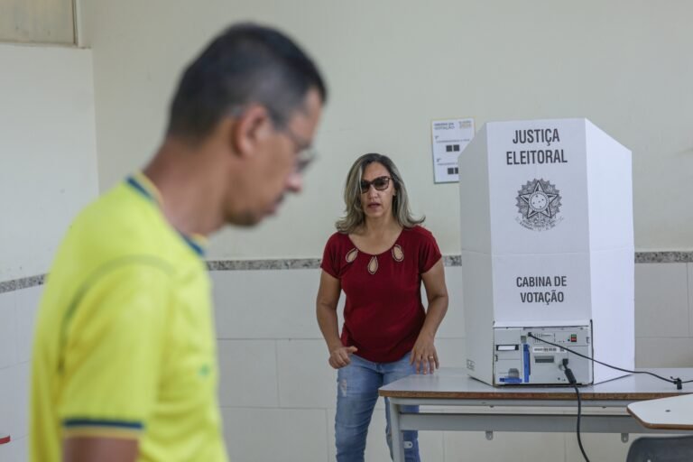 votos de Lula: Eleitora aguarda ao lado de urna eletrônica com cabine da Justiça Eleitoral durante votação no segundo turno de 2026.