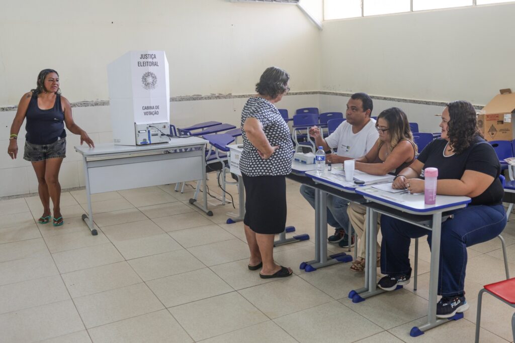 transporte para eleitores: Eleitores e mesários em uma seção eleitoral com urna eletrônica e cabine de votação.