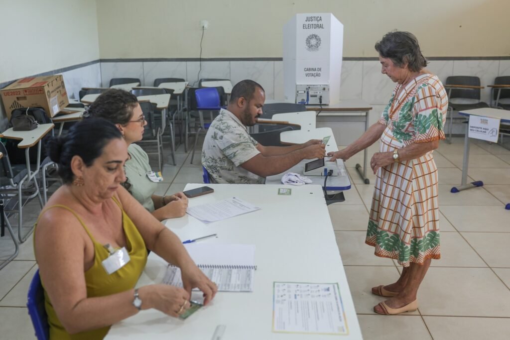 venda de voto: Idosa vota em seção eleitoral com urna eletrônica diante de mesários em sala de aula.