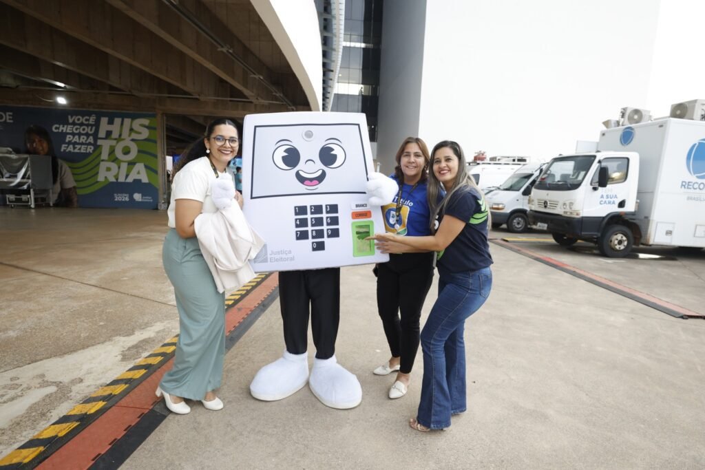 horario de votacao: Três mulheres sorrindo ao lado de uma pessoa fantasiada de urna eletrônica, com um painel da Justiça Eleitoral ao fundo.